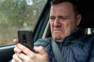 Sad millennial guy looks at phone screen in surprise, sits in passenger seat. Disgusted and overwhel