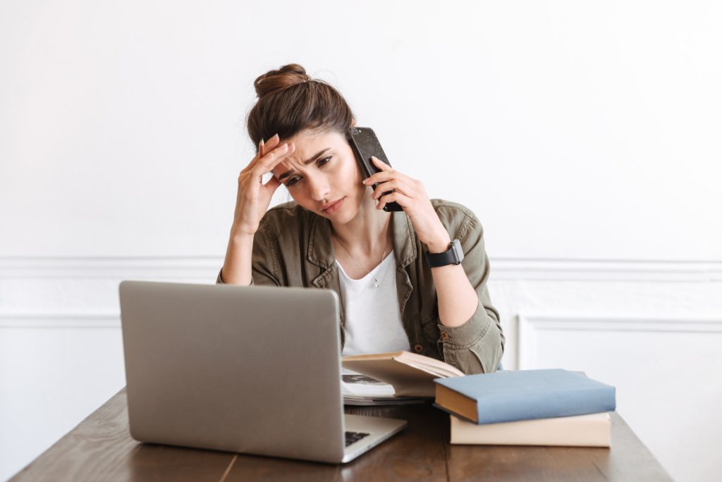 Young confused displeased woman using laptop computer indoors talking by mobile phone.