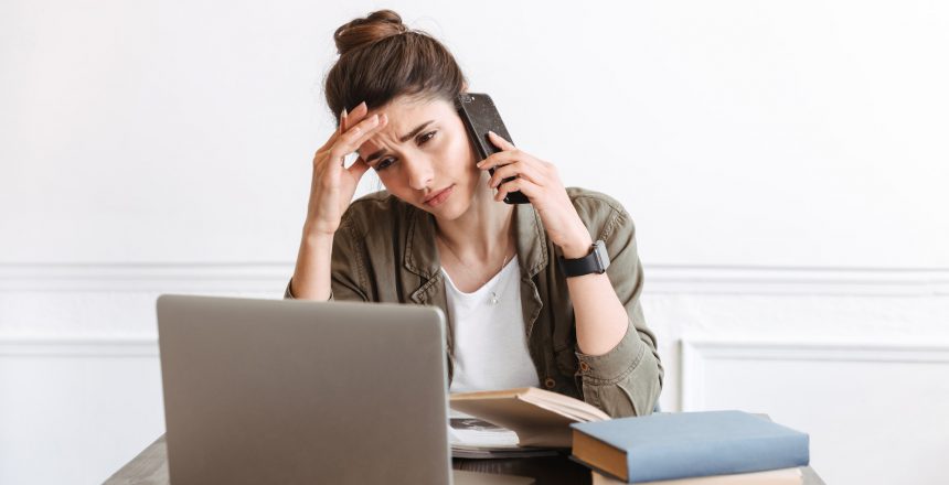 Young confused displeased woman using laptop computer indoors talking by mobile phone.